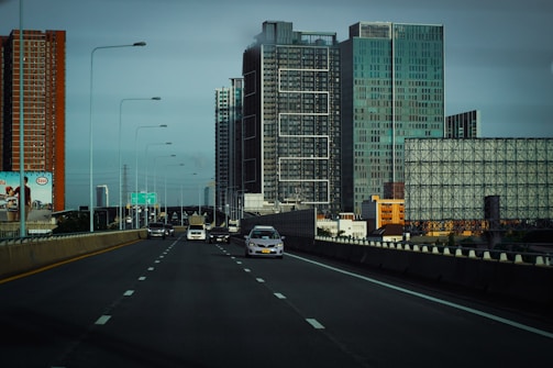 A highway scene with multiple vehicles, including cars and a truck, driving on a multi-lane road. Skyscrapers and tall buildings of various designs are visible in the background, with a clear sky overhead. A billboard with an advertisement is positioned on the side of the road.