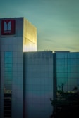 A modern building with reflective glass windows and a large red sign displaying the Isuzu logo. The structure is highlighted by light, creating a contrast between the illuminated portions and the surrounding areas. The sky provides a serene backdrop with subtle shades of blue and green.