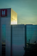 A modern building with reflective glass windows and a large red sign displaying the Isuzu logo. The structure is highlighted by light, creating a contrast between the illuminated portions and the surrounding areas. The sky provides a serene backdrop with subtle shades of blue and green.