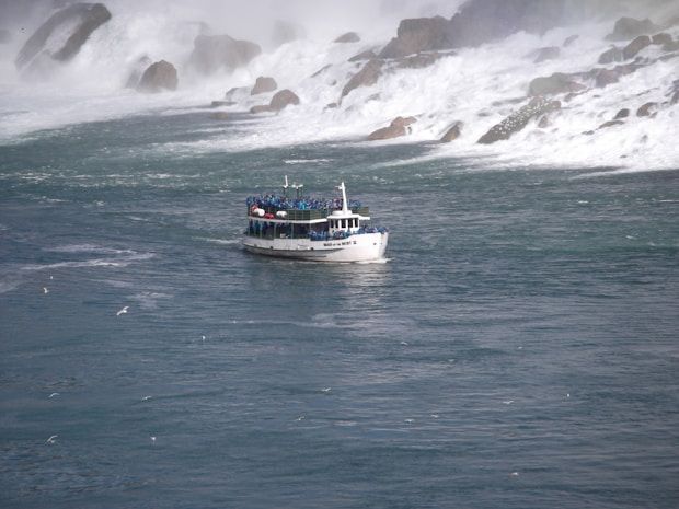 A tour boat filled with passengers sails on a wide river near cascading waterfalls. The water is turbulent, and mist rises from where the falls crash into the river, creating a dramatic backdrop. Seagulls fly around the scene, adding to the sense of motion and activity.