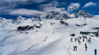 A snow-covered mountainous landscape with a group of people engaging in outdoor winter activities. The sky is partly cloudy, and ski lifts can be seen in the distance, suggesting a ski resort location.