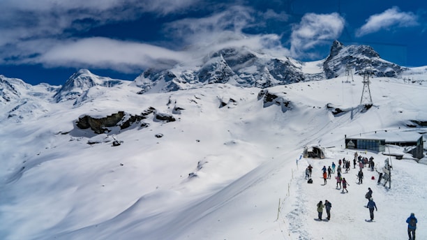 A snow-covered mountainous landscape with a group of people engaging in outdoor winter activities. The sky is partly cloudy, and ski lifts can be seen in the distance, suggesting a ski resort location.