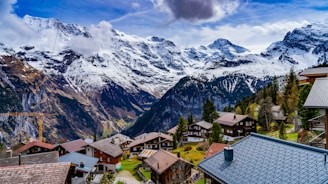 Charming alpine village nestled in a lush valley with snow-capped mountains in the background. Traditional wooden houses with sloped roofs are scattered throughout the scene, surrounded by evergreens and vibrant green grass. A clear blue sky with scattered clouds hangs above the majestic peaks.