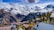 Charming alpine village nestled in a lush valley with snow-capped mountains in the background. Traditional wooden houses with sloped roofs are scattered throughout the scene, surrounded by evergreens and vibrant green grass. A clear blue sky with scattered clouds hangs above the majestic peaks.