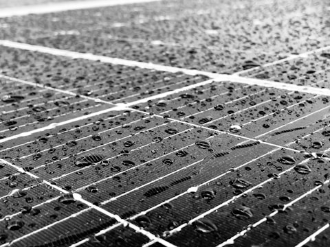 Close-up of a solar panel surface covered in numerous raindrops. The texture is visible, with droplets of varying sizes reflecting light. The panel is composed of multiple cells with visible grid lines that create a geometric pattern across the surface.
