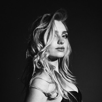 A black and white photograph featuring a woman with long, wavy hair looking off into the distance. Her expression is calm and confident. The lighting creates strong contrasts, highlighting her facial features and hair. The composition is elegant and artistic.