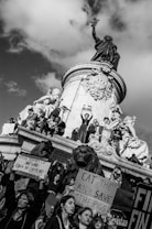 A group of people gather around and atop a large monument, holding signs with messages in support of nature and against economic inequality. The monument features statues of figures and a large lion. The atmosphere is one of activism and protest, with visible expressions of determination among the participants.