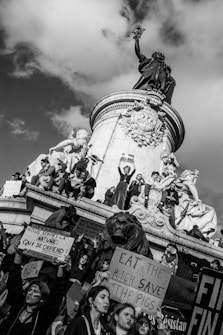 A group of people gather around and atop a large monument, holding signs with messages in support of nature and against economic inequality. The monument features statues of figures and a large lion. The atmosphere is one of activism and protest, with visible expressions of determination among the participants.