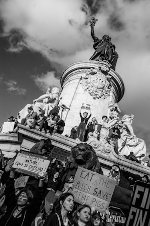 A group of people gather around and atop a large monument, holding signs with messages in support of nature and against economic inequality. The monument features statues of figures and a large lion. The atmosphere is one of activism and protest, with visible expressions of determination among the participants.