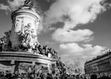 A large statue depicts a seated figure surrounded by people, some holding banners. The sky is partly cloudy, and there are classic European buildings in the background.