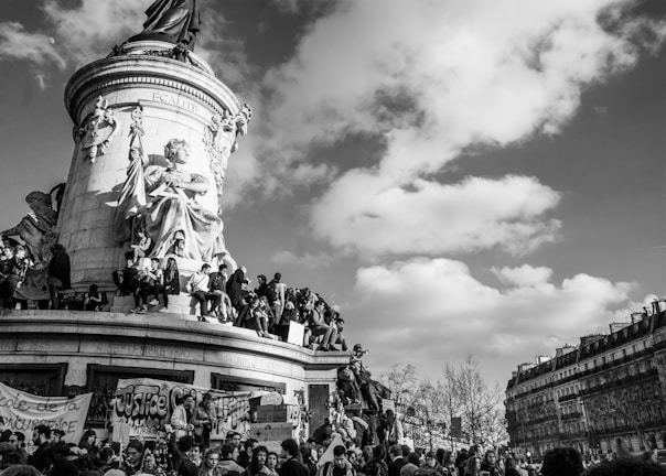 A large statue depicts a seated figure surrounded by people, some holding banners. The sky is partly cloudy, and there are classic European buildings in the background.