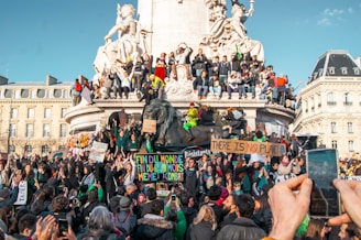 A large group of people are gathered in front of a monumental sculpture. Many individuals are holding banners with messages about climate change. Some people have climbed onto the statue, which features a lion and seated figures. The crowd appears engaged and energetic, as some capture the moment with their phones.