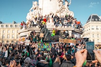 A large group of people are gathered in front of a monumental sculpture. Many individuals are holding banners with messages about climate change. Some people have climbed onto the statue, which features a lion and seated figures. The crowd appears engaged and energetic, as some capture the moment with their phones.