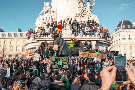 A large group of people are gathered in front of a monumental sculpture. Many individuals are holding banners with messages about climate change. Some people have climbed onto the statue, which features a lion and seated figures. The crowd appears engaged and energetic, as some capture the moment with their phones.