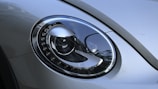 A close-up of a modern car's headlight featuring a sleek, oval design with intricate detailing around the edges. The surface is reflective and smooth, displaying the surrounding environment faintly.