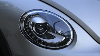 A close-up of a modern car's headlight featuring a sleek, oval design with intricate detailing around the edges. The surface is reflective and smooth, displaying the surrounding environment faintly.