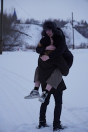 A smiling family ski sherpa carrying multiple pairs of skis and boots through fresh snow.