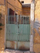 A rustic gate featuring faded green paint with visible rust patches stands against an aged, yellow ochre wall. The gate is flanked by stone columns with peeling plaster, and there are electrical wires running above. A number plate with the number 35 is affixed to the wall.