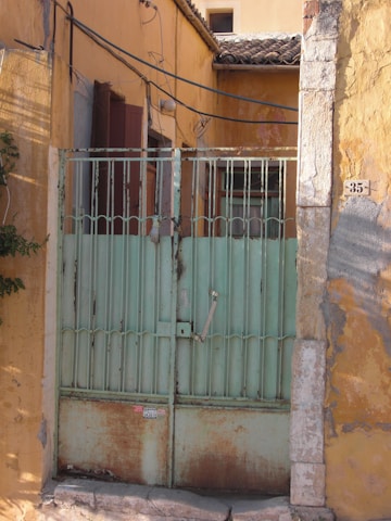 A rustic gate featuring faded green paint with visible rust patches stands against an aged, yellow ochre wall. The gate is flanked by stone columns with peeling plaster, and there are electrical wires running above. A number plate with the number 35 is affixed to the wall.