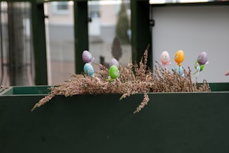 Decorative Easter eggs are placed on sticks and inserted into a bed of dried heather plants in a green planter. The background is blurred, showing parts of a building or structure.