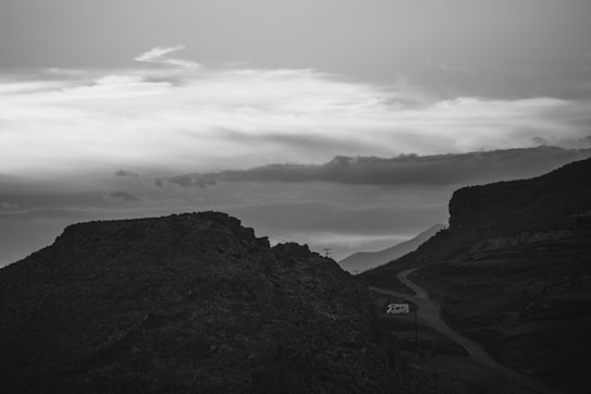 A black and white landscape depicting a rugged mountain terrain with a winding road leading through it. A camper van is parked on the side of the road, surrounded by the rocky environment. Above, a vast sky is filled with layered clouds, creating a dramatic contrast against the silhouetted landscape.