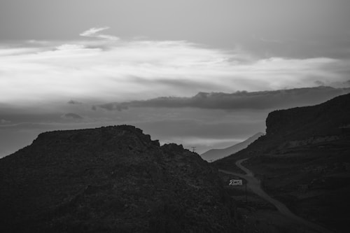 A black and white landscape depicting a rugged mountain terrain with a winding road leading through it. A camper van is parked on the side of the road, surrounded by the rocky environment. Above, a vast sky is filled with layered clouds, creating a dramatic contrast against the silhouetted landscape.