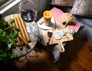 A cozy setting with a wooden table featuring a glass of red wine, a wooden cutting board with blue cheese, and bread on a red checkered cloth. A lit candle adds warmth to the scene, next to neatly arranged honey-colored candles and a green plant.
