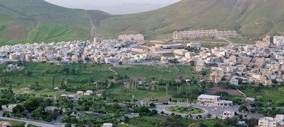A densely packed cityscape is nestled between rolling green hills. The area is predominantly residential, with numerous buildings tightly clustered together. In the foreground, roads and a roundabout enhance the urban setting, surrounded by lush greenery.