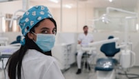 A dental professional is wearing a mask and a blue surgical cap adorned with dental-themed prints. She is looking back towards the camera in a dental clinic setting. Another person in a white uniform is seated in the background near dental equipment. The room is bright and clean, featuring dental chairs and tools.