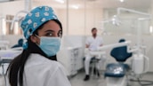 A dental professional is wearing a mask and a blue surgical cap adorned with dental-themed prints. She is looking back towards the camera in a dental clinic setting. Another person in a white uniform is seated in the background near dental equipment. The room is bright and clean, featuring dental chairs and tools.