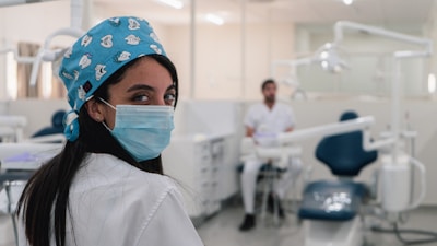 A dental professional is wearing a mask and a blue surgical cap adorned with dental-themed prints. She is looking back towards the camera in a dental clinic setting. Another person in a white uniform is seated in the background near dental equipment. The room is bright and clean, featuring dental chairs and tools.
