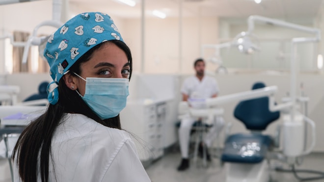 A dental professional is wearing a mask and a blue surgical cap adorned with dental-themed prints. She is looking back towards the camera in a dental clinic setting. Another person in a white uniform is seated in the background near dental equipment. The room is bright and clean, featuring dental chairs and tools.