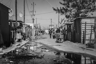 A black and white street scene in an informal settlement with makeshift houses constructed from corrugated metal. People are seen walking and standing on the street, and a person is pushing a shopping cart. Power lines and poles are visible across the skyline, and there is litter strewn across the ground with puddles reflecting the surroundings.