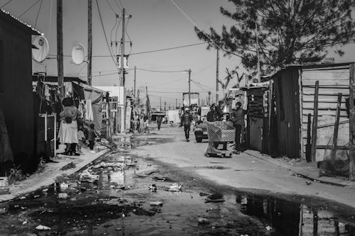 A black and white street scene in an informal settlement with makeshift houses constructed from corrugated metal. People are seen walking and standing on the street, and a person is pushing a shopping cart. Power lines and poles are visible across the skyline, and there is litter strewn across the ground with puddles reflecting the surroundings.