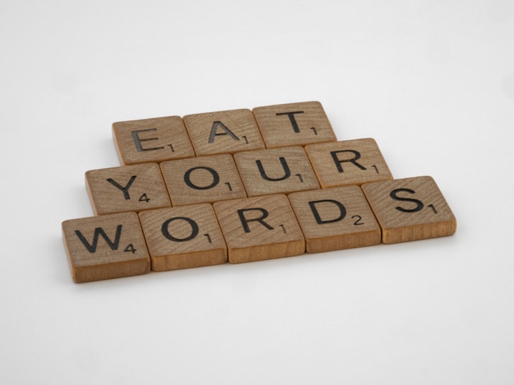 Wooden letter tiles arranged to form the words 'EAT YOUR WORDS'. Each tile includes a letter and a corresponding point value typically used in word games.