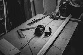 A woodworking table with various tools and wooden pieces. A smoothing plane and a set square are placed alongside planks of wood. A person's hands rest on the table as they work on the wooden project. Ear protection headphones are also visible.
