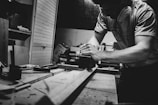 A black and white image of a person working intently in a workshop. The individual is sanding a piece of wood on a workbench surrounded by tools. The setting appears to be a woodworking shop with various materials and equipment visible in the background.