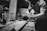 A black and white image of a person working intently in a workshop. The individual is sanding a piece of wood on a workbench surrounded by tools. The setting appears to be a woodworking shop with various materials and equipment visible in the background.