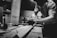 A black and white image of a person working intently in a workshop. The individual is sanding a piece of wood on a workbench surrounded by tools. The setting appears to be a woodworking shop with various materials and equipment visible in the background.