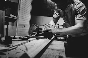 A black and white image of a person working intently in a workshop. The individual is sanding a piece of wood on a workbench surrounded by tools. The setting appears to be a woodworking shop with various materials and equipment visible in the background.