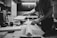 A person is working in a woodworking shop, handling a piece of wood on a workbench surrounded by tools such as clamps and hearing protection earmuffs. The setting appears organized and focused on carpentry or woodworking tasks.