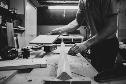 A person is working in a woodworking shop, handling a piece of wood on a workbench surrounded by tools such as clamps and hearing protection earmuffs. The setting appears organized and focused on carpentry or woodworking tasks.