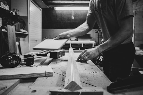 A person is working in a woodworking shop, handling a piece of wood on a workbench surrounded by tools such as clamps and hearing protection earmuffs. The setting appears organized and focused on carpentry or woodworking tasks.