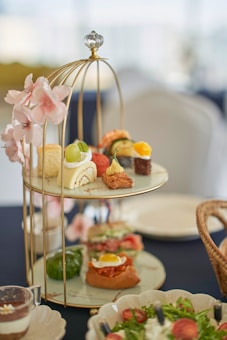 A multi-tiered serving stand displays an assortment of elegant finger foods and desserts. The top tier features small pastries and a roll cake garnished with fruit. Pink floral decorations adorn the stand, adding a touch of elegance. In the foreground, there are plates of fresh salad and a cappuccino glass.