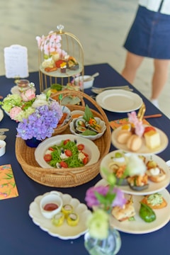 A variety of beautifully presented dishes are set on a dark blue tablecloth. There are plates with a vibrant green salad garnished with cherry tomatoes and cheese, a basket with leafy greens, and a multi-tiered stand displaying small bites and desserts. Floral arrangements with purple and light pink flowers add a decorative touch. A person is partially visible in the background.