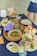 A variety of beautifully presented dishes are set on a dark blue tablecloth. There are plates with a vibrant green salad garnished with cherry tomatoes and cheese, a basket with leafy greens, and a multi-tiered stand displaying small bites and desserts. Floral arrangements with purple and light pink flowers add a decorative touch. A person is partially visible in the background.