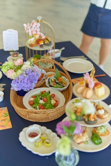 A variety of beautifully presented dishes are set on a dark blue tablecloth. There are plates with a vibrant green salad garnished with cherry tomatoes and cheese, a basket with leafy greens, and a multi-tiered stand displaying small bites and desserts. Floral arrangements with purple and light pink flowers add a decorative touch. A person is partially visible in the background.