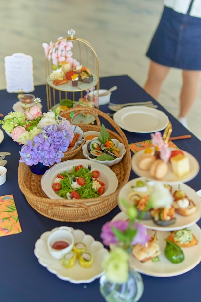A variety of beautifully presented dishes are set on a dark blue tablecloth. There are plates with a vibrant green salad garnished with cherry tomatoes and cheese, a basket with leafy greens, and a multi-tiered stand displaying small bites and desserts. Floral arrangements with purple and light pink flowers add a decorative touch. A person is partially visible in the background.