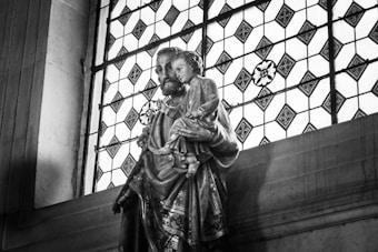 A religious statue depicting a bearded man holding a child is placed in front of a detailed stained glass window. The window features geometric patterns and floral designs, adding to the sacred atmosphere.