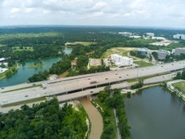 An aerial view of a highway running through a landscape featuring green trees, water bodies, and several buildings. The highway is busy with multiple lanes of traffic. Surrounding areas include a lush forest, ponds, and office buildings with parking areas. The sky appears cloudy, contributing to an overcast atmosphere.